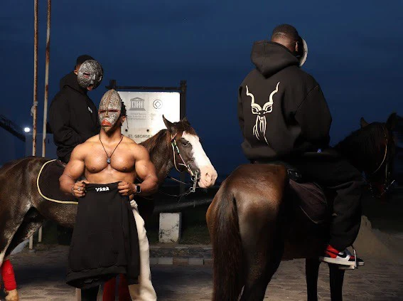 Three figures in black Yárà outerwear at dusk beside two horses, one hoodie carrying the white kudu emblem across the back.